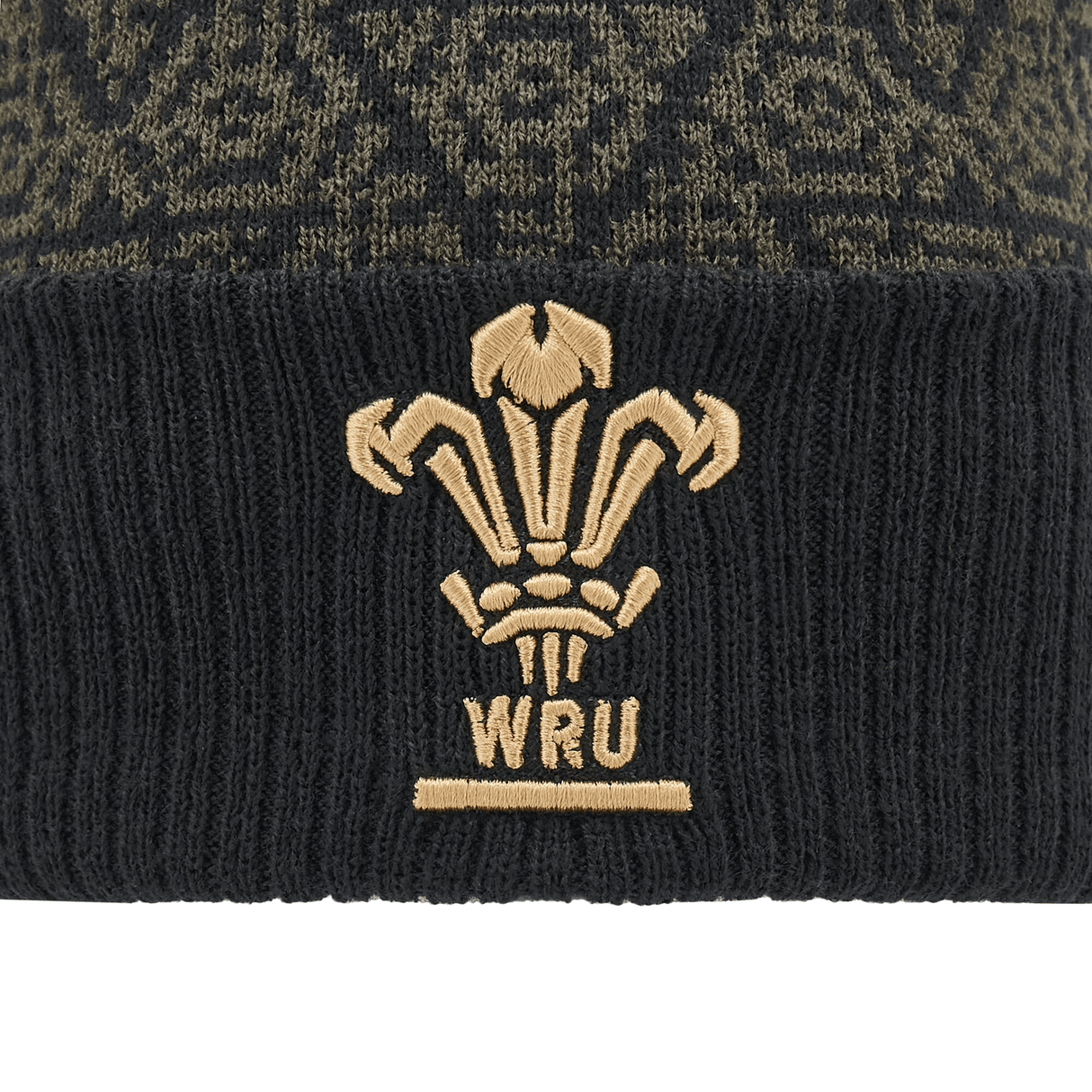 Close-up of the Wales 24/25 Black Pom Pom Beanie by Macron, featuring a gold emblem with three feathers and "WRU" embroidered below.