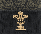 Close-up of the Wales 24/25 Black Pom Pom Beanie by Macron, featuring a gold emblem with three feathers and "WRU" embroidered below.
