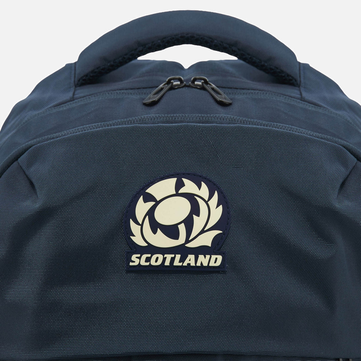 Close-up of the Scotland SRU 26/27 Backpack by Macron in blue, showcasing a prominent Scotland Rugby emblem patch centered on the front pocket. The backpack features a top carry handle and two visible zipper pulls—making it the perfect sports backpack for loyal Scotland rugby supporters.