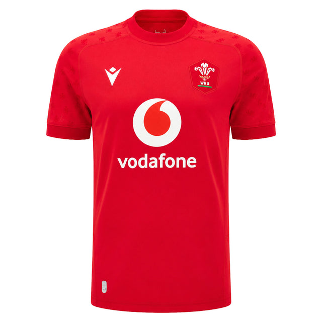 The Wales WRU 26/27 Replica Home Jersey by Macron is a red short-sleeve rugby shirt featuring the white Vodafone logo, WRU crest, and Macron branding on the front. Part of the official Macron 26/27 collection, this replica jersey is perfect for supporters looking to show their pride.
