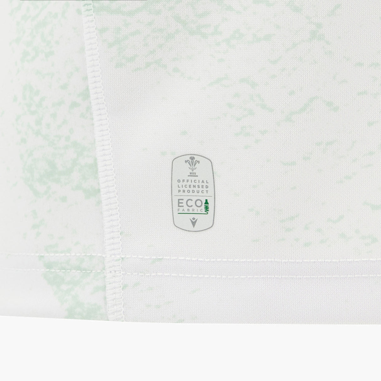 Close-up of the Wales WRU 26/27 Replica Away Jersey by Macron, featuring a white base with a light green pattern and a tag displaying “Official Licensed Product Eco Fabric” alongside a small tree icon.