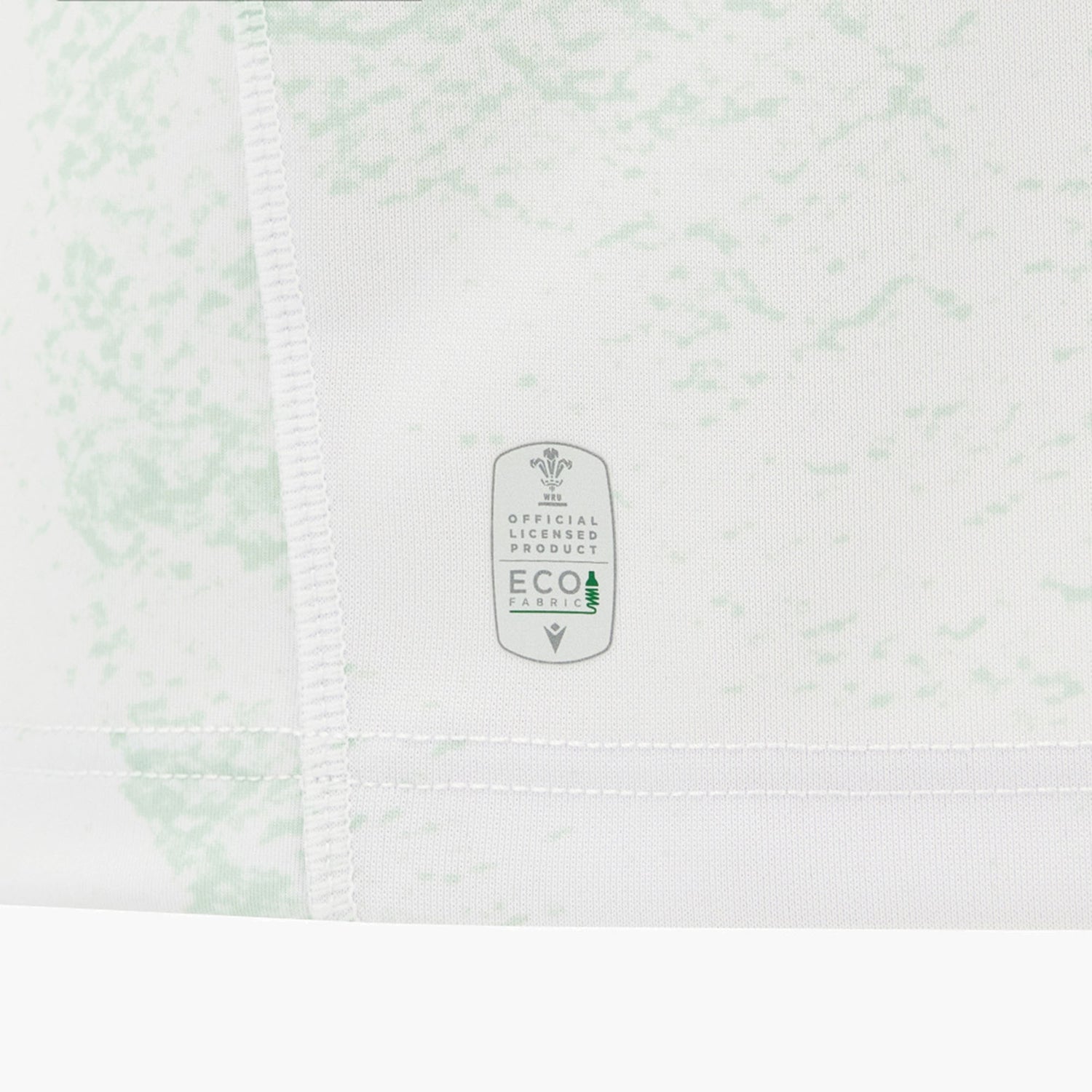Close-up of the Wales WRU 26/27 Replica Away Jersey by Macron, featuring a white base with a light green pattern and a tag displaying “Official Licensed Product Eco Fabric” alongside a small tree icon.