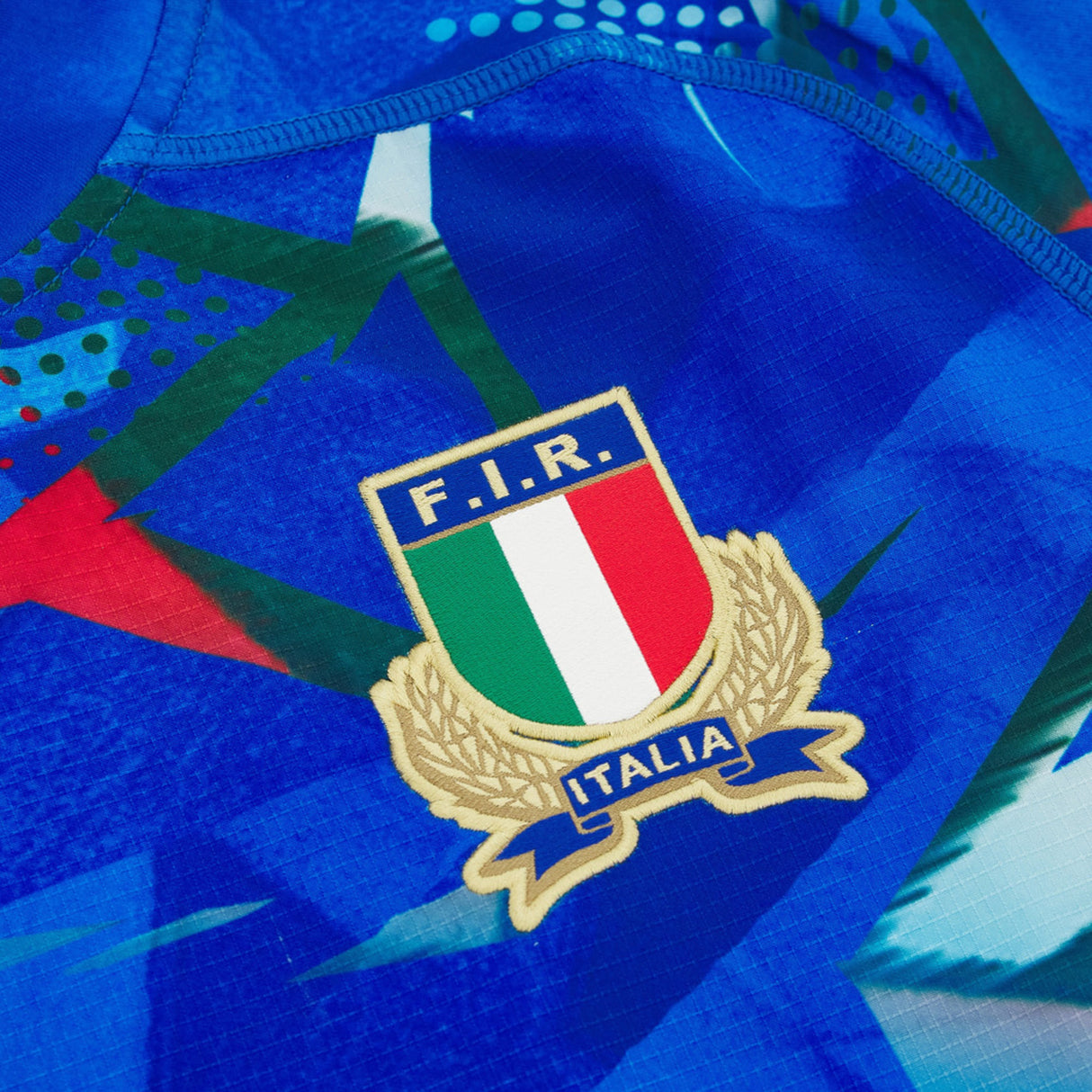 Close-up of the Italy 26/27 Home Authentic Jersey by Macron, featuring the F.I.R. (Federazione Italiana Rugby) crest with an Italian flag and gold laurel wreath on a blue patterned fabric background—an official Macron jersey that proudly represents Italy’s national heritage and sporting excellence.