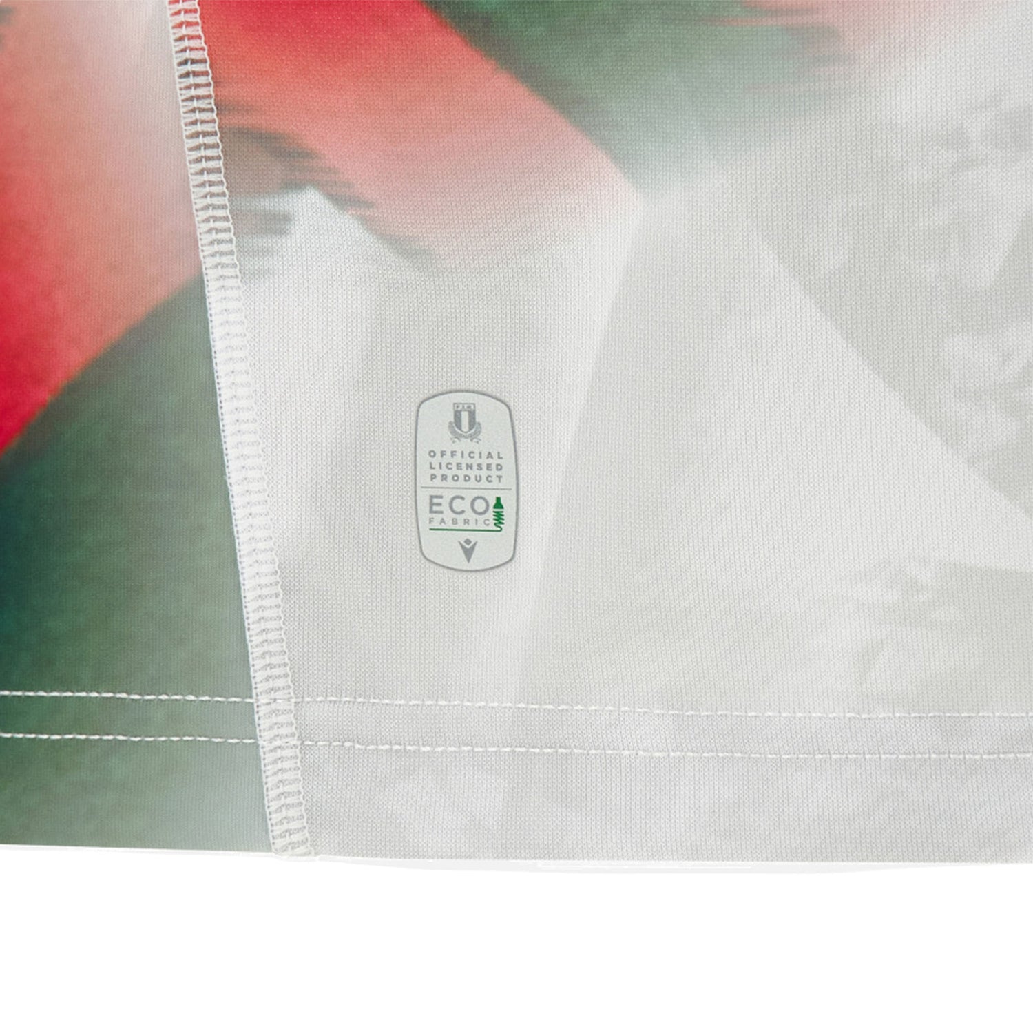 Close-up of a fabric featuring a white label that reads “Official Product ECO Fabric” with two green trees, stitched onto the Italy 26/27 Away Replica Jersey by Macron, which displays an abstract red, green, and white pattern.