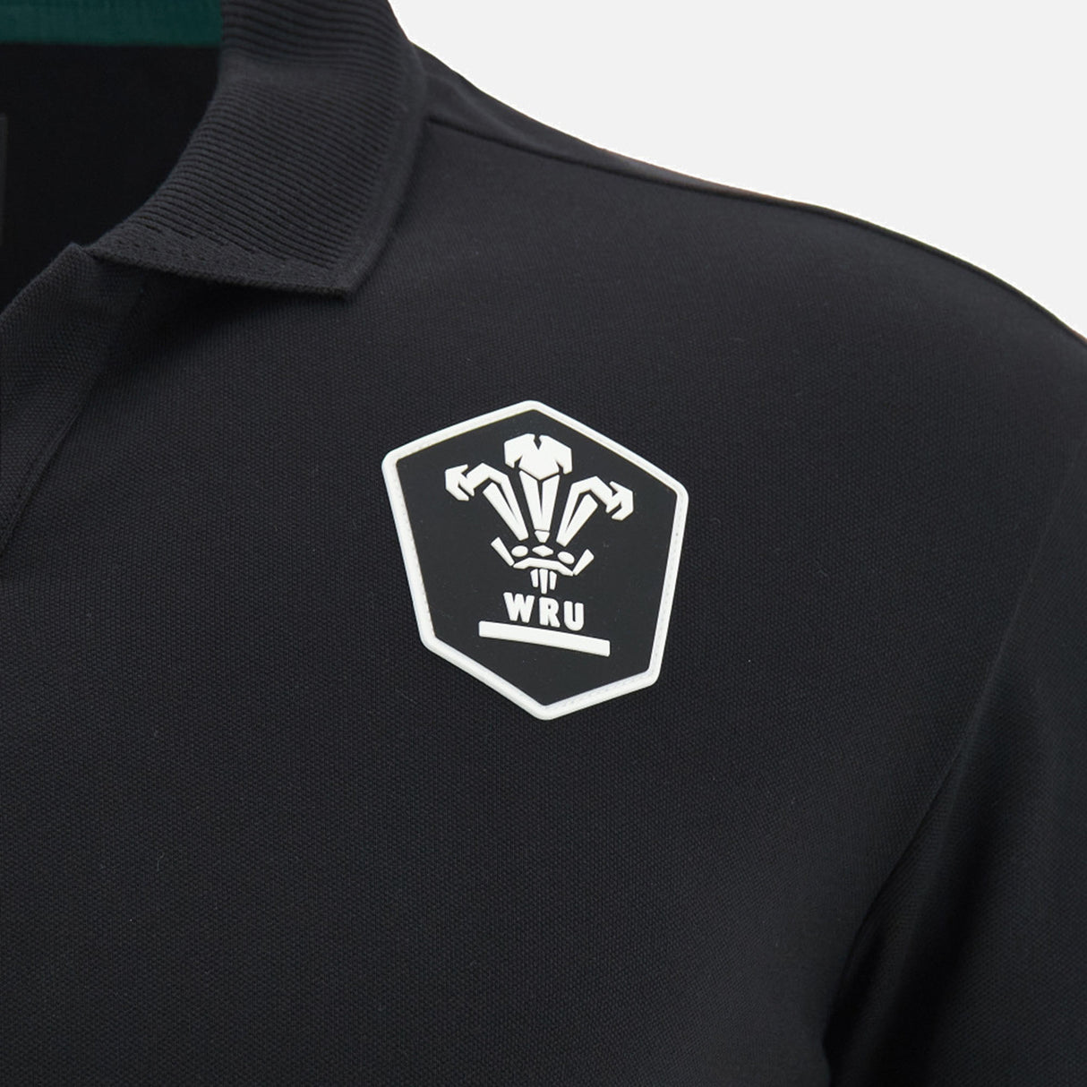 The Wales WRU 26/27 Travel Cotton Polo by Macron is a black polo shirt styled like a classic Macron rugby top, featuring a white WRU logo with three feathers inside a shield on the left chest.