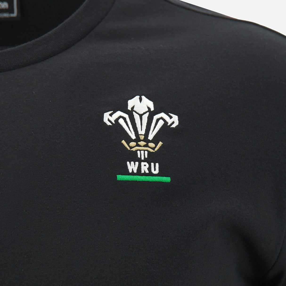 Discover the Wales WRU 26/27 Travel Poly/Cotton T-Shirt by Macron, shown here in black as part of the official WRU travel collection. This stylish t-shirt features the Welsh Rugby Union (WRU) logo expertly embroidered in white, gold, and green on the chest, complemented by iconic Macron branding for an added touch of quality craftsmanship.