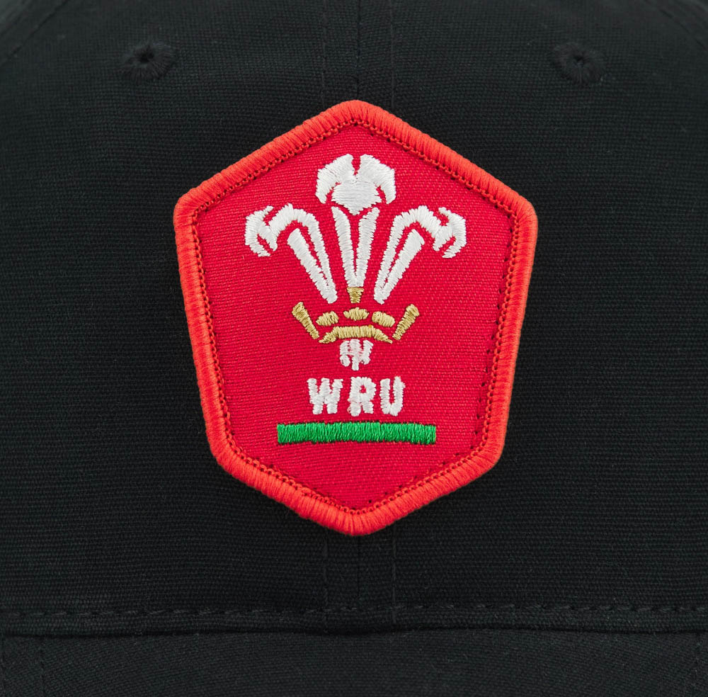 Wales WRU 26/27 Dad Cap by Macron—a black cap featuring a red pentagonal patch with the Welsh Rugby Union (WRU) logo, white three-feather emblem, and green and gold accents; a classic piece from Macron ideal for fans seeking both style and heritage.
