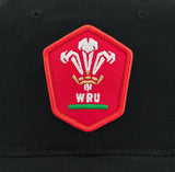 Wales WRU 26/27 Dad Cap by Macron—a black cap featuring a red pentagonal patch with the Welsh Rugby Union (WRU) logo, white three-feather emblem, and green and gold accents; a classic piece from Macron ideal for fans seeking both style and heritage.