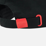 Close-up of a black fabric strap with a red plastic buckle and red loop, reminiscent of the detailing found on the Wales WRU 26/27 Dad Cap by Macron. The buckle features the embossed text “GYMRU.” The background is plain light gray.