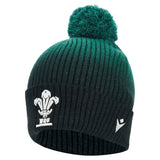 The Wales WRU 26/27 Pom-Pom Beanie by Macron is a ribbed knit green and black beanie featuring a pom-pom on top and embroidered white Macron logos on the front and side.