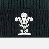 A close-up of the Wales WRU 26/27 Pom-Pom Beanie by Macron, a black ribbed knit beanie featuring the white embroidered Welsh Rugby Union (WRU) logo.