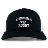 The EMB S&S Activewear Birmingham Rugby Text Logo Retro Trucker is a black and white hat featuring “Birmingham Rugby” text and a small anvil logo embroidered on the front. This cap offers a classic snapback closure for an adjustable fit.