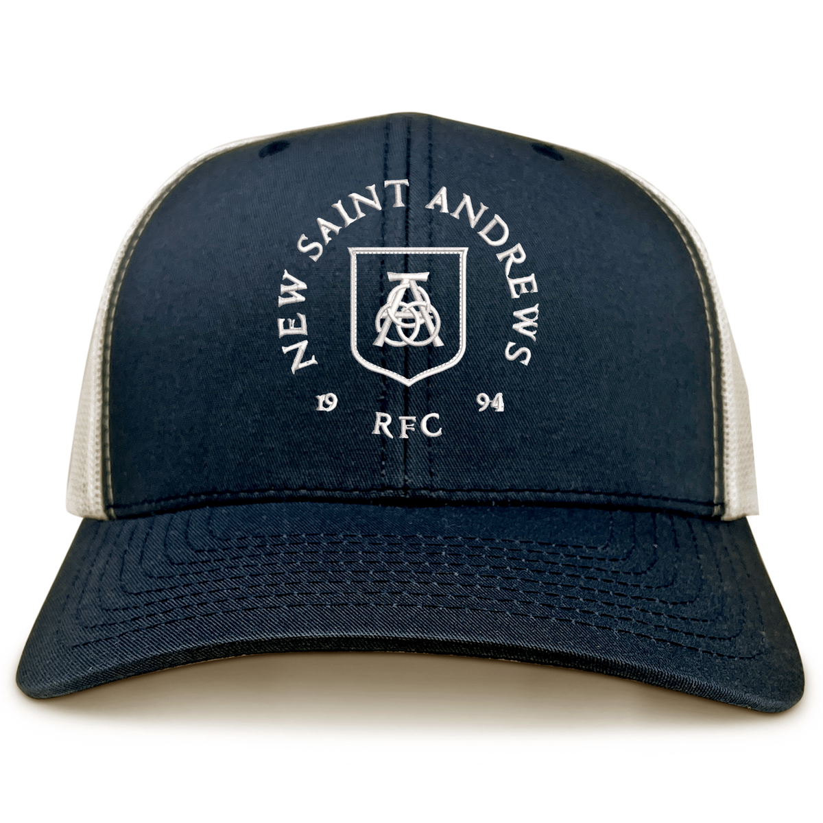 The New Saint Andrews College Retro Trucker Cap by EMB S&S Activewear is a navy blue and white hat featuring an embroidered “New Saint Andrews RFC 1994” shield logo on the front, complete with a comfortable snapback closure.