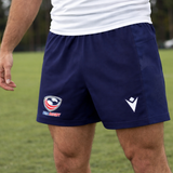 A person wearing USA Rugby 25/26 Matchday Home Shorts by Macron in navy blue and a white shirt stands on a grassy field.