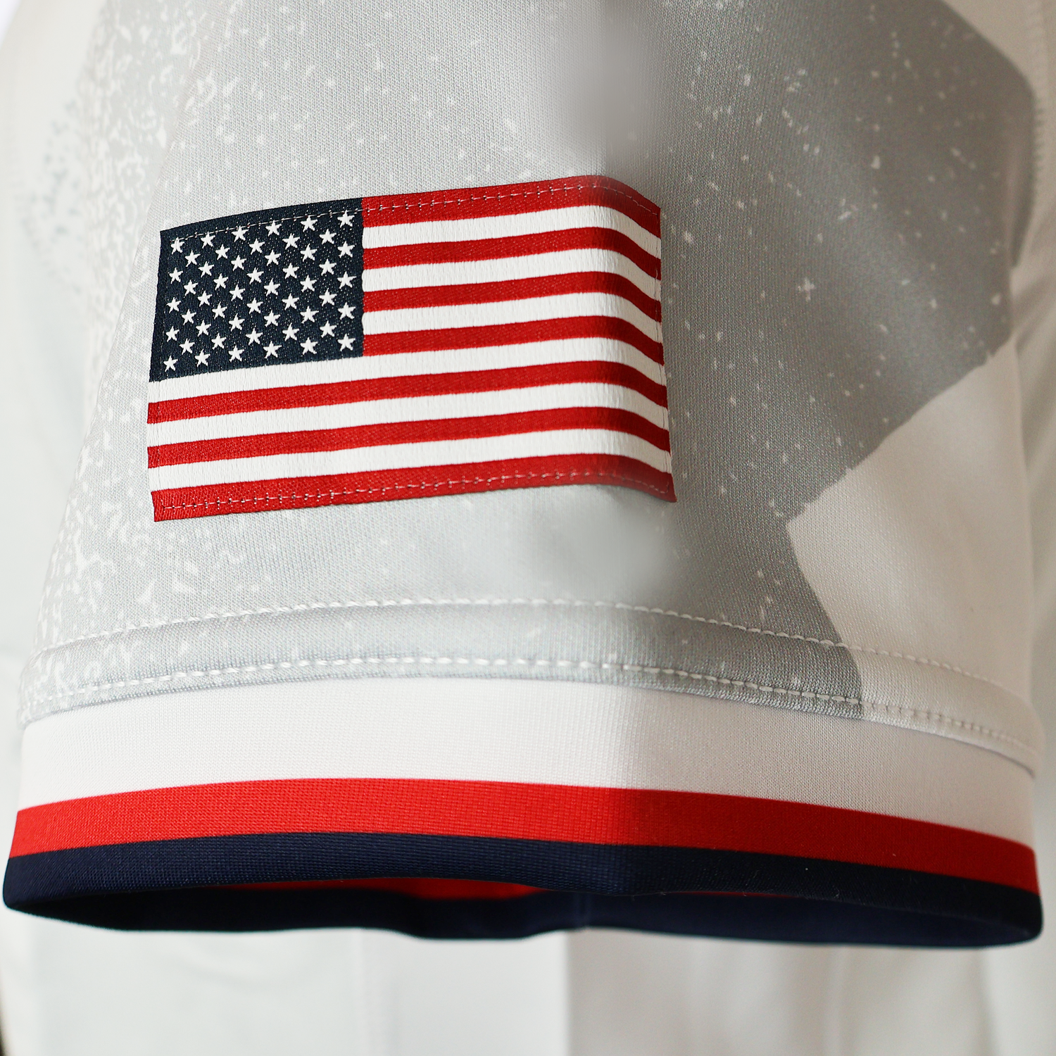 A close-up of an American flag patch sewn onto a white garment with red, white, and blue trim from the USA Rugby 24/25 Authentic Matchday Away Jersey by Macron.