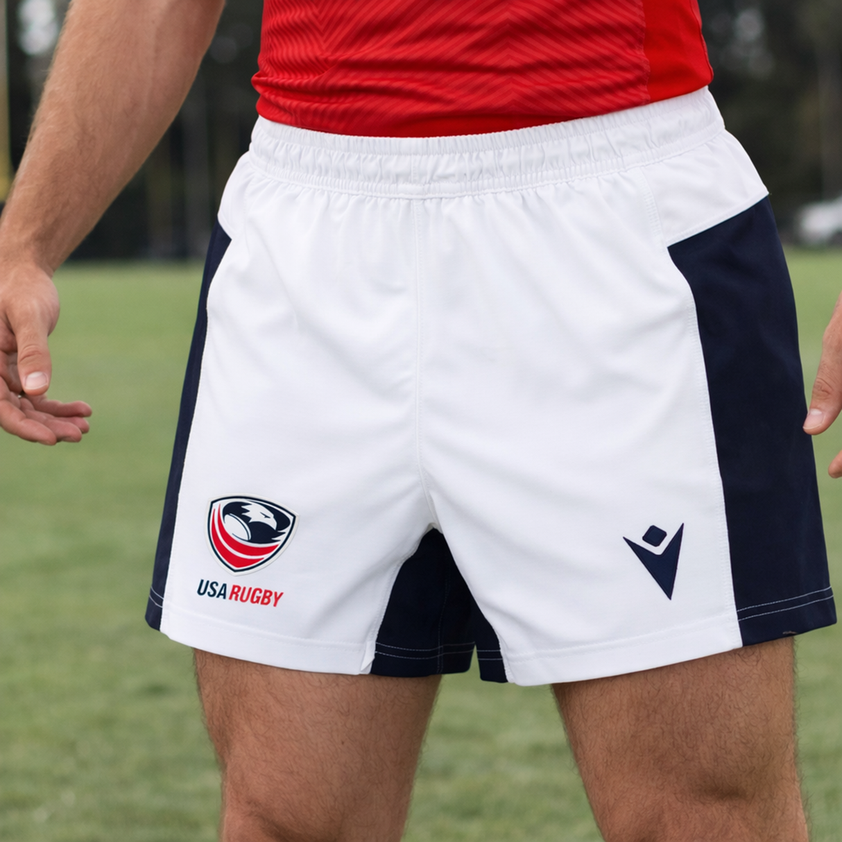 A person in USA Rugby 24/25 Matchday Away Shorts by Macron—white shorts with navy side panels—and a red shirt stands on a grassy field.
