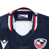 Close-up of the collar and chest of the Women's USA Rugby 24/25 Home Bodyfit Shirt by Macron, in navy with red, white, and blue trim, featuring a bird logo and the Macron brand label.