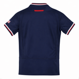 The Youth USA Rugby 24/25 Home Jersey by Macron is a navy short-sleeve rugby shirt viewed from the back, with red, white, and blue trim on the collar/sleeves and a small American flag patch on the left sleeve for classic USA Rugby style.