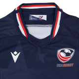 The Youth USA Rugby 24/25 Home Jersey by Macron is a navy blue jersey featuring the USA Rugby eagle logo, the Macron logo, and red, white, and blue collar accents.