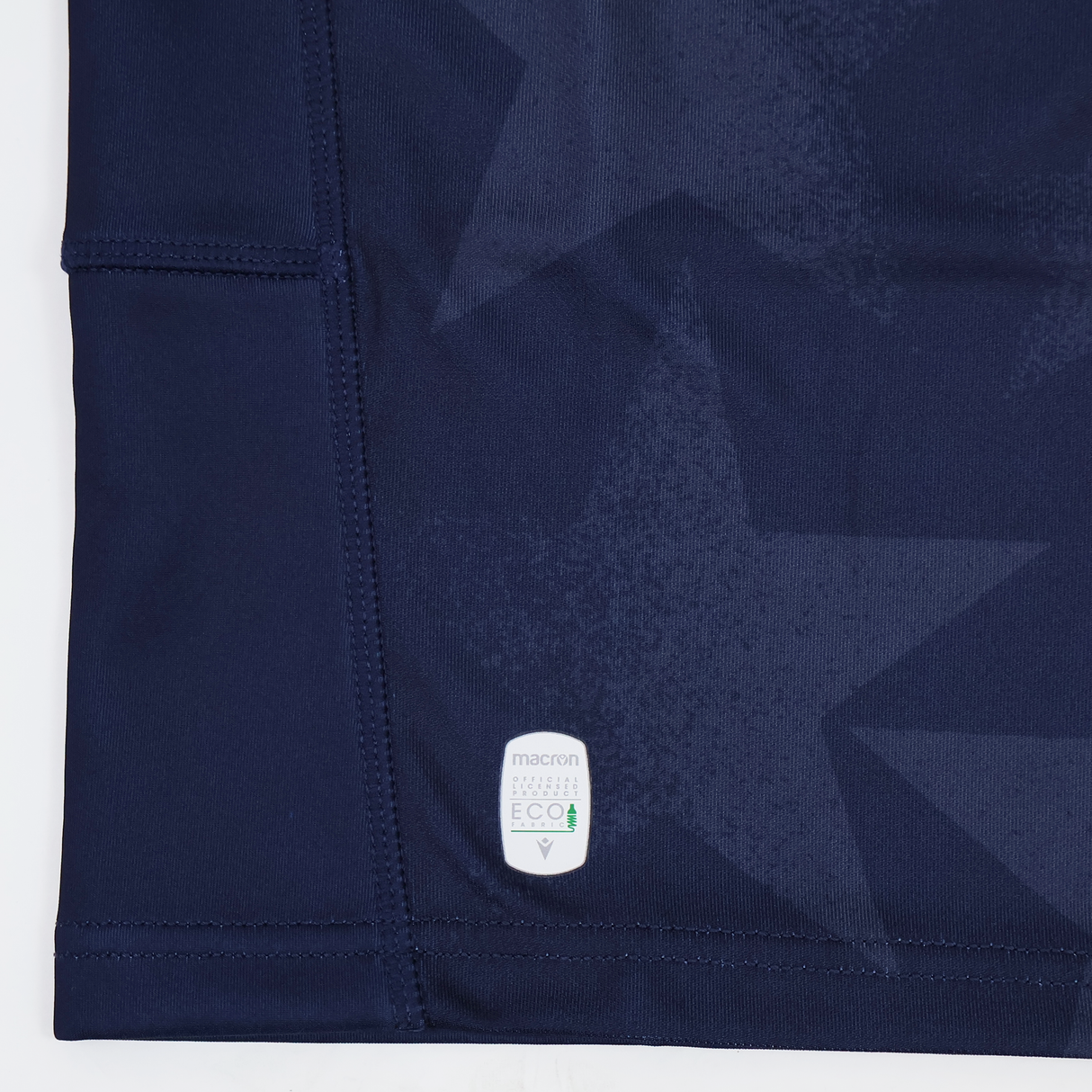 Close-up of navy blue fabric with subtle star patterns, visible stitching, and a small white "macron ECO" label near the hem—part of the Youth USA Rugby 24/25 Home Jersey by Macron.