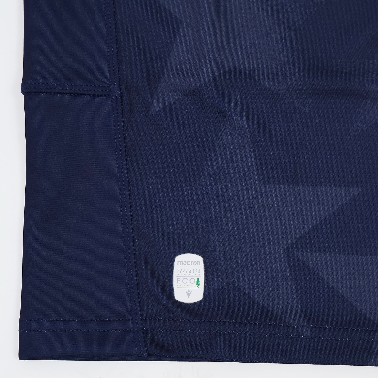 Close-up of navy blue fabric with subtle star patterns, visible stitching, and a small white "macron ECO" label near the hem—part of the Youth USA Rugby 24/25 Home Jersey by Macron.