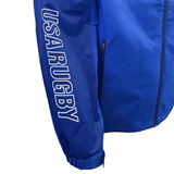The USA Rugby 25 Player Waterproof Jacket by Macron is blue, has a side zip pocket, and "USA RUGBY" printed in white on the left sleeve.