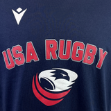 The USA Rugby 25 Leisure T-Shirt by Macron in navy blue features "USA Rugby" in bold red outlined letters with a white and red eagle logo underneath.