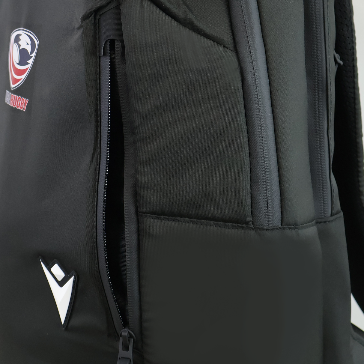 Close-up of the USA Rugby 25/26 Staff Backpack by Macron in black, showing a partially unzipped side pocket, rugby logo, and a white geometric emblem on the front.