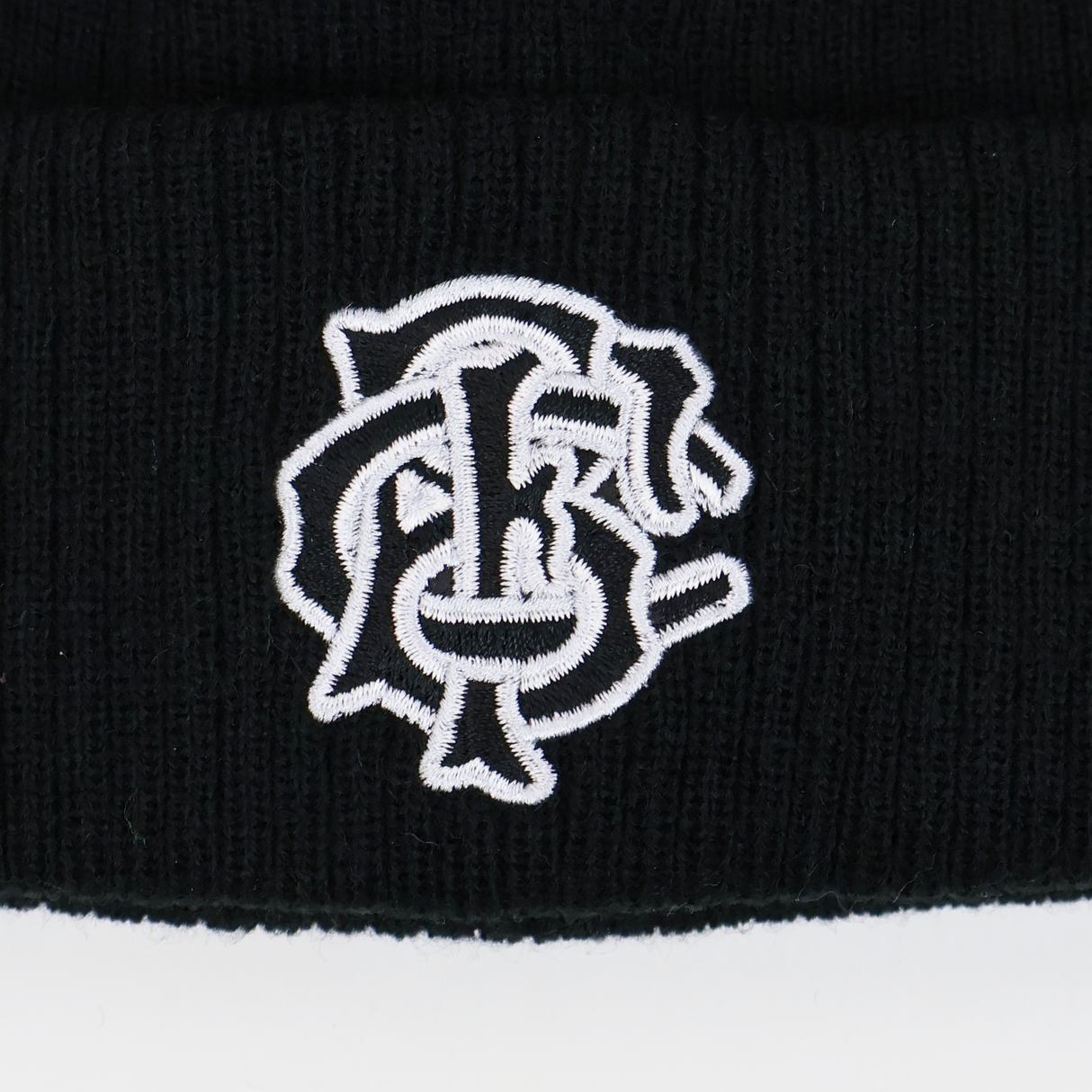 A close-up of the black Barbarians 25/26 Pom-Pom Beanie by Macron, featuring a white embroidered monogram logo with intertwined letters.