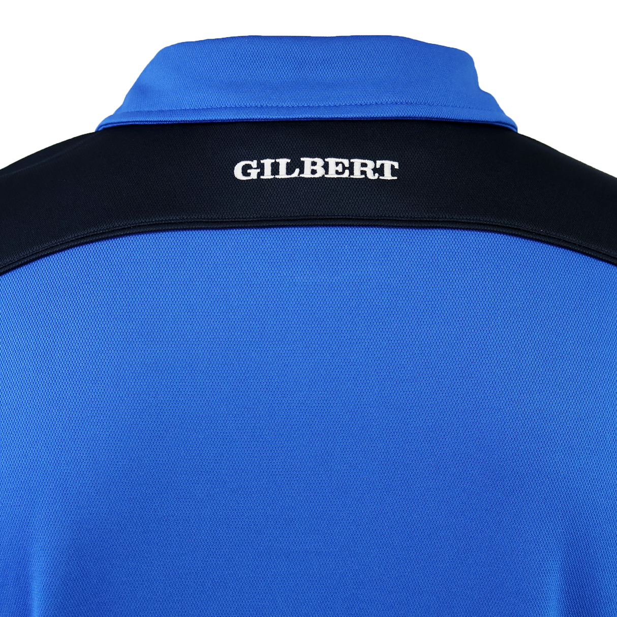 Close-up of the back of a blue Gilbert Photon II Polo with a black yoke, lightweight fabric, and "GILBERT" embroidered in white on the upper back.
