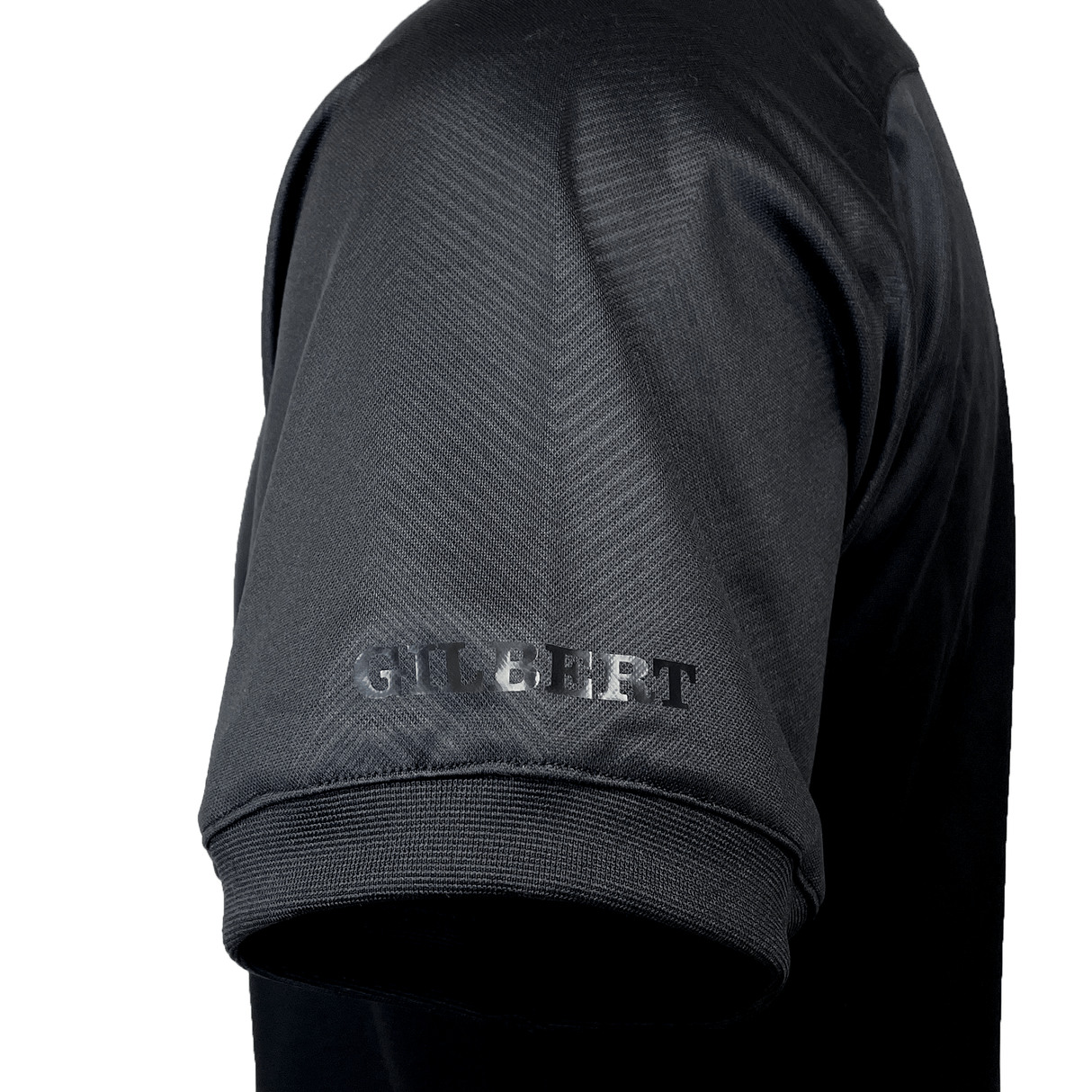 Close-up of the left sleeve of a black short-sleeved Gilbert Evo Performance Training Tee with "GILBERT" written in gray text above the hem.