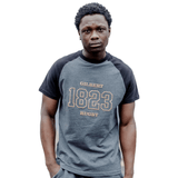A person stands facing the camera, wearing a dark gray Gilbert Rugby 200 Year Anniversary Leisure Shirt by Gilbert with "Gilbert Rugby 1823" printed on the front, celebrating nearly 200 years of rugby history.