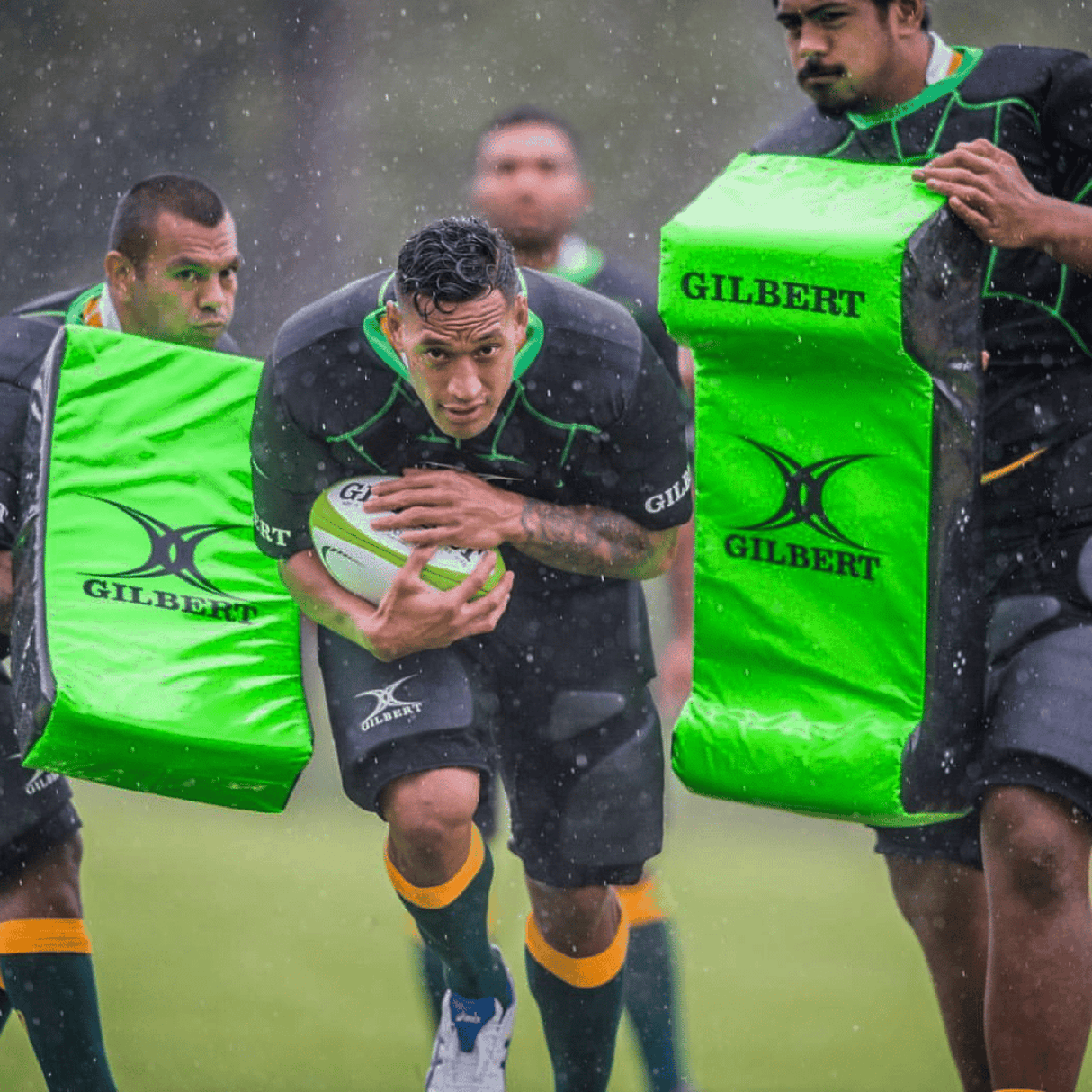 Three rugby players practice in the rain; one runs with the ball while two others, wearing padded Gilbert Contact Top Pro shirts, hold bright green tackling pads labeled "GILBERT.