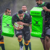 Three rugby players practice in the rain; one runs with the ball while two others, wearing padded Gilbert Contact Top Pro shirts, hold bright green tackling pads labeled "GILBERT.