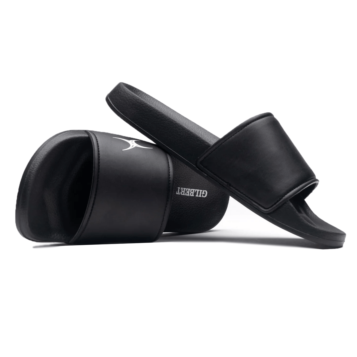 Two black Gilbert Slide Sandals are displayed; one standing upright and the other lying on its side, showcasing the brand name "Gilbert" on the sole. Crafted from quick-drying material with plush cushioning, they offer both comfort and convenience.