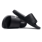 Two black Gilbert Slide Sandals are displayed; one standing upright and the other lying on its side, showcasing the brand name "Gilbert" on the sole. Crafted from quick-drying material with plush cushioning, they offer both comfort and convenience.