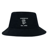 The EMB S&S Activewear Amoskeag Rugby Club Crusher Bucket Cap is a black cotton twill hat with white "AMOSKEAG," an illustrated ram’s head, and "Est. 1984" embroidery—stylish sun protection for any occasion.