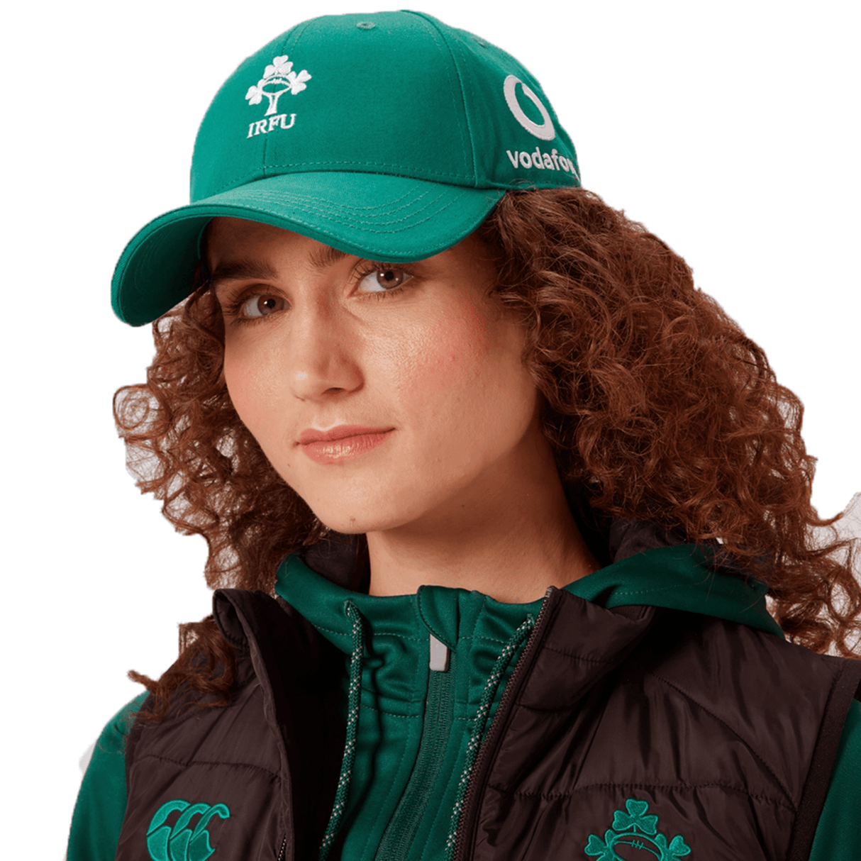 A person with curly hair is wearing a green Ireland 24 Drill Cap by Canterbury featuring high definition Ireland logo embroidery, along with a hooded jacket and a black vest.