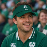 A man in an Ireland rugby jersey and a Canterbury Ireland 26 Visor smiles while sitting in a stadium with blurred fans in the background.