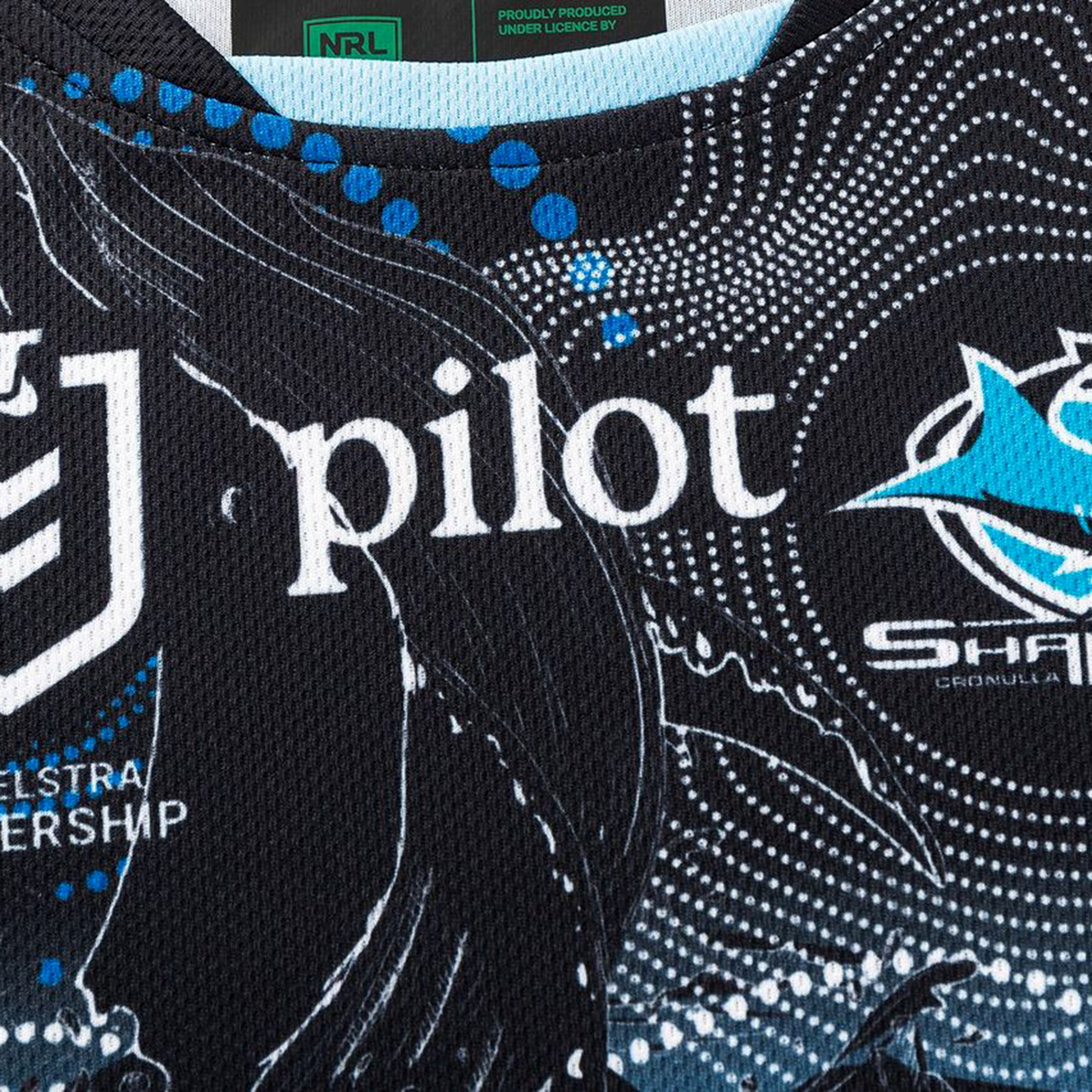 Close-up of the Sharks Indigenous NRL 25/26 Replica Jersey by Classic Sportswear, showing the Telstra NRL Premiership, "pilot" sponsor logos, and part of the Cronulla Sharks logo in white and blue on black patterned fabric.