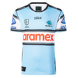 The Sharks NRL 26/27 Replica Home Jersey by Classic Sportswear is light blue with black, white, and blue stripes, featuring the Cronulla Sharks logo, NRL Premiership logo, and "aramex" sponsor across the chest.