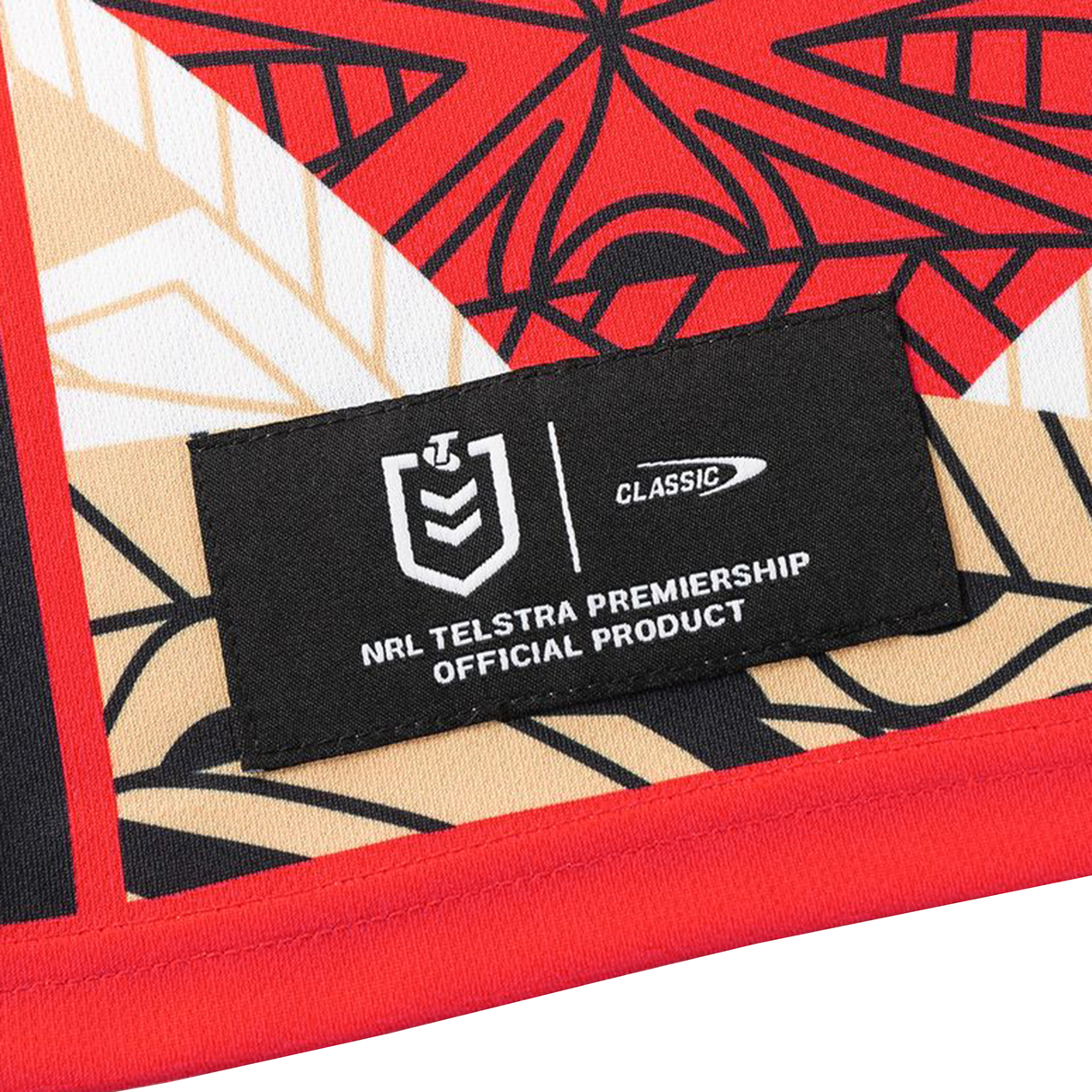 A fabric label reading "NRL Telstra Premiership Official Product" is attached to the Dolphins Indigenous NRL 25/26 Replica Jersey by Classic Sportswear, featuring a geometric red, black, beige, and white pattern.