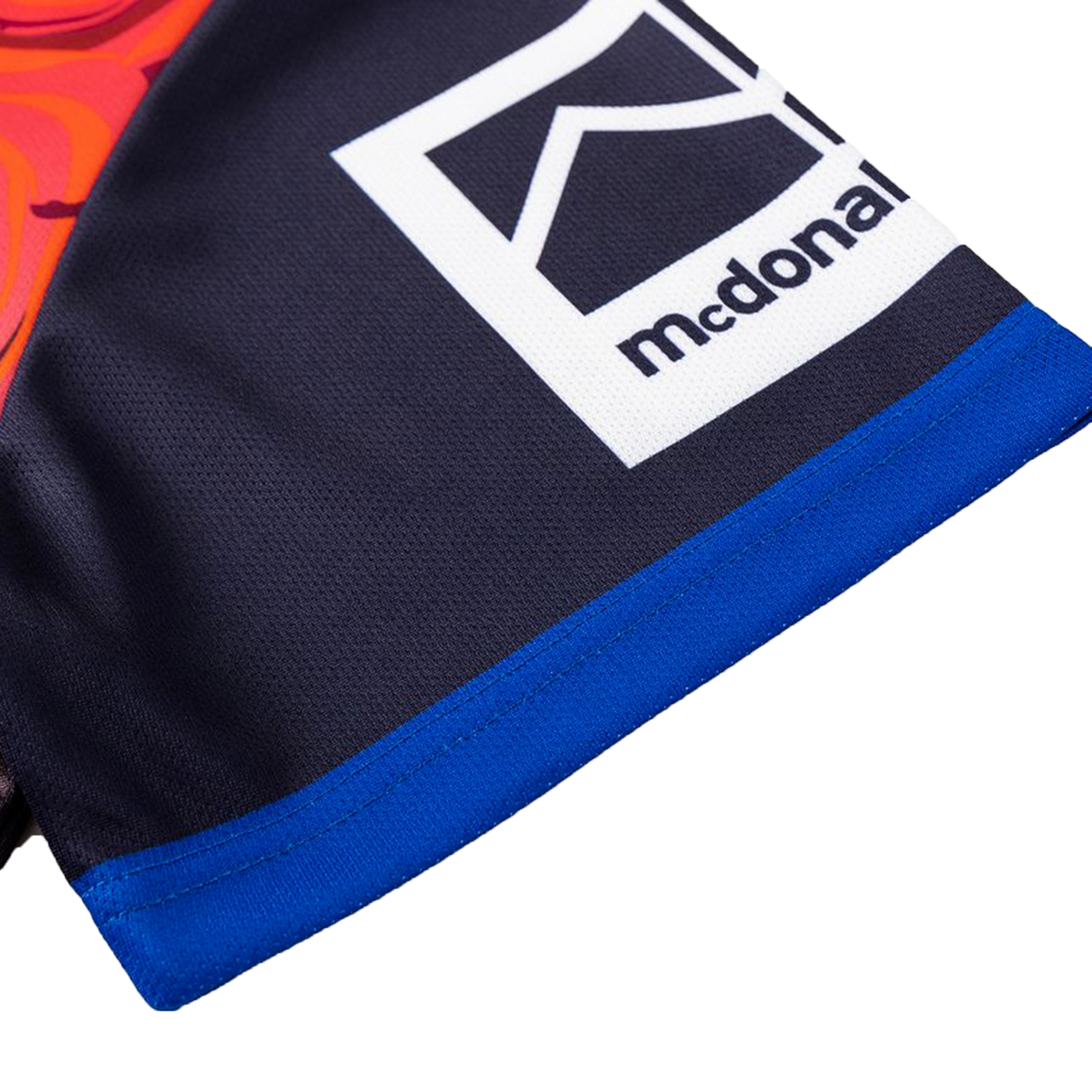 Close-up of a garment with a blue hem and a rectangular patch displaying a stylized house icon and "mcdonal," inspired by the Knights Indigenous NRL 25/26 Replica Jersey from Classic Sportswear.