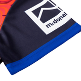 Close-up of a garment with a blue hem and a rectangular patch displaying a stylized house icon and "mcdonal," inspired by the Knights Indigenous NRL 25/26 Replica Jersey from Classic Sportswear.