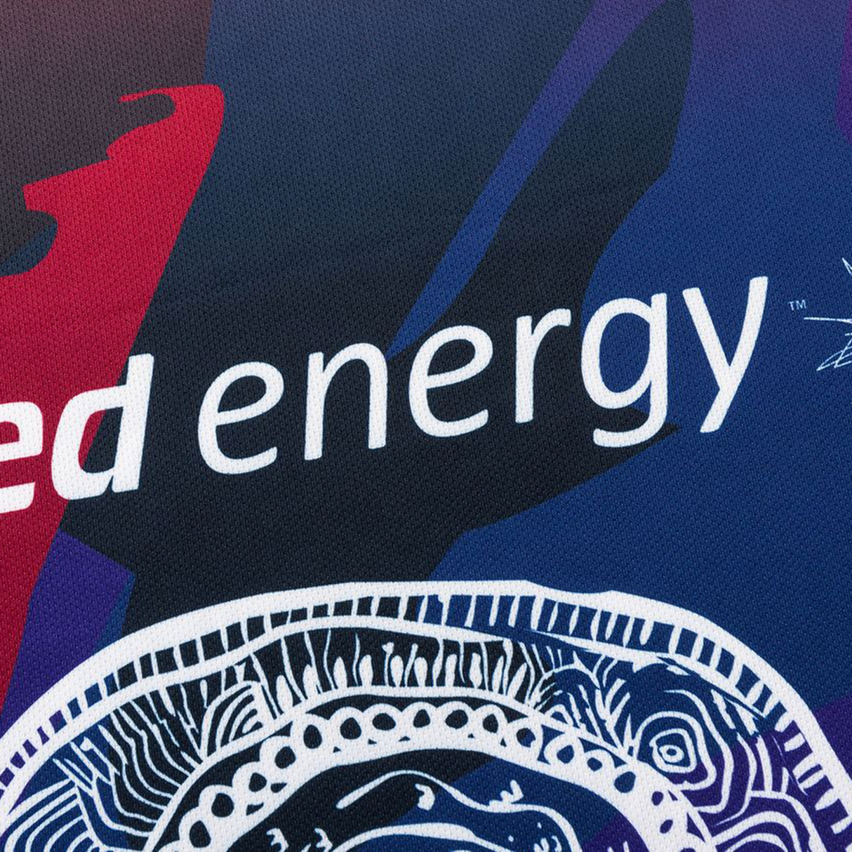 Close-up of fabric from the Knights Indigenous NRL 25/26 Replica Jersey by Classic Sportswear, featuring abstract blue, red, and white patterns with partially visible “ed energy” text.