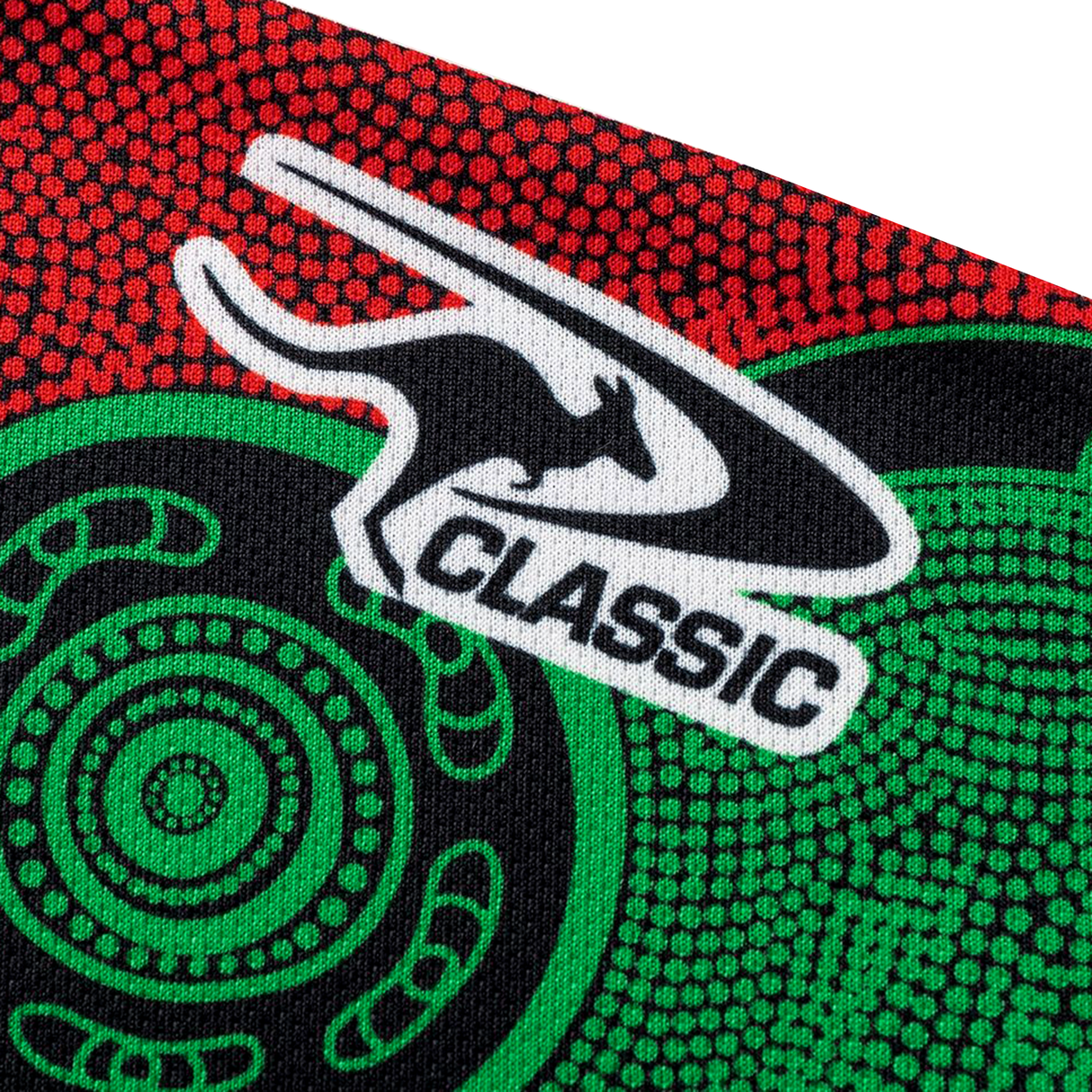 Close-up of fabric with a black kangaroo logo and "CLASSIC" on a red and green patterned background, showcasing the Classic Sportswear Rabbitohs Indigenous NRL 25/26 Replica Jersey.