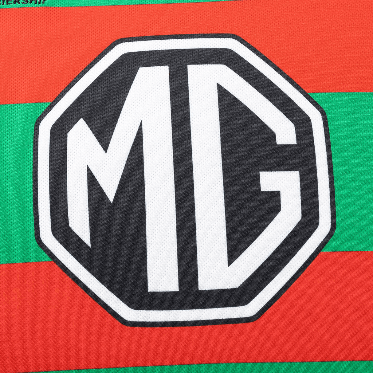 Close-up of the Classic Sportswear logo with "MG" in bold white outlined in black, set against red and green stripes emulating a Rabbitohs NRL 25/26 Replica Home Jersey by Classic Sportswear.