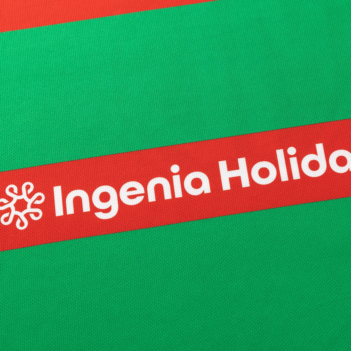 Close-up of a red and green fabric with "Ingenia Holiday" and a star logo on the red stripe, from the Rabbitohs NRL 26/27 Replica Home Jersey by Classic Sportswear.