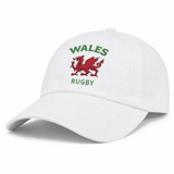 The EMB S&S Activewear Nations of Rugby Wales Dad Cap is a white cotton cap featuring a red Welsh dragon and green "WALES RUGBY" embroidery—a classic choice for rugby fans.