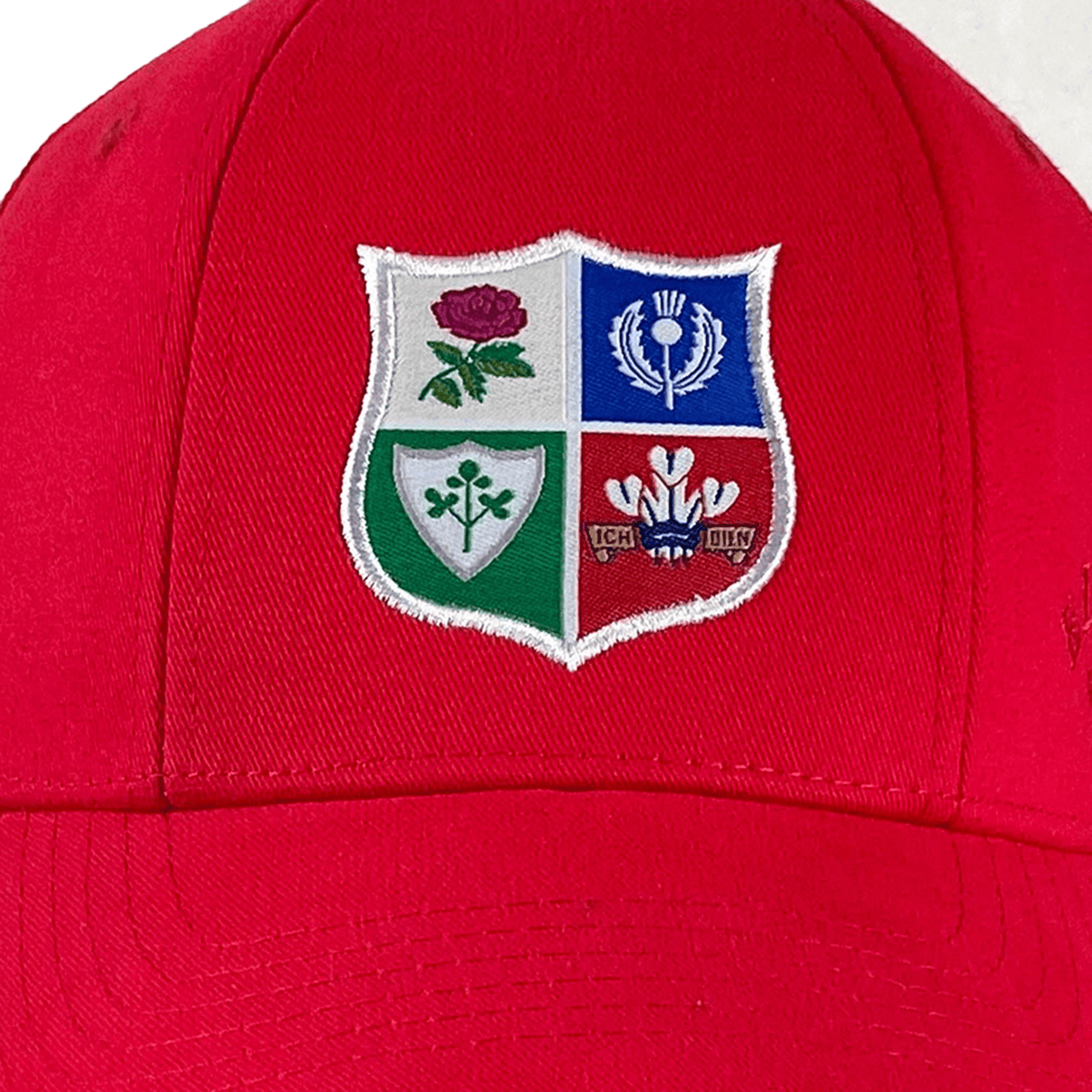 The Ellis Rugby British Rugby 1971 Lions Tour Cap features a red design with an embroidered emblem in four quadrants, showcasing rose, thistle, shamrock, and feather symbols. It celebrates rugby heritage as a homage by Ellis Rugby.